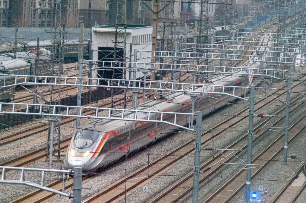 Ein silberner Zug fährt auf Gleisen neben hohen Gebäuden.